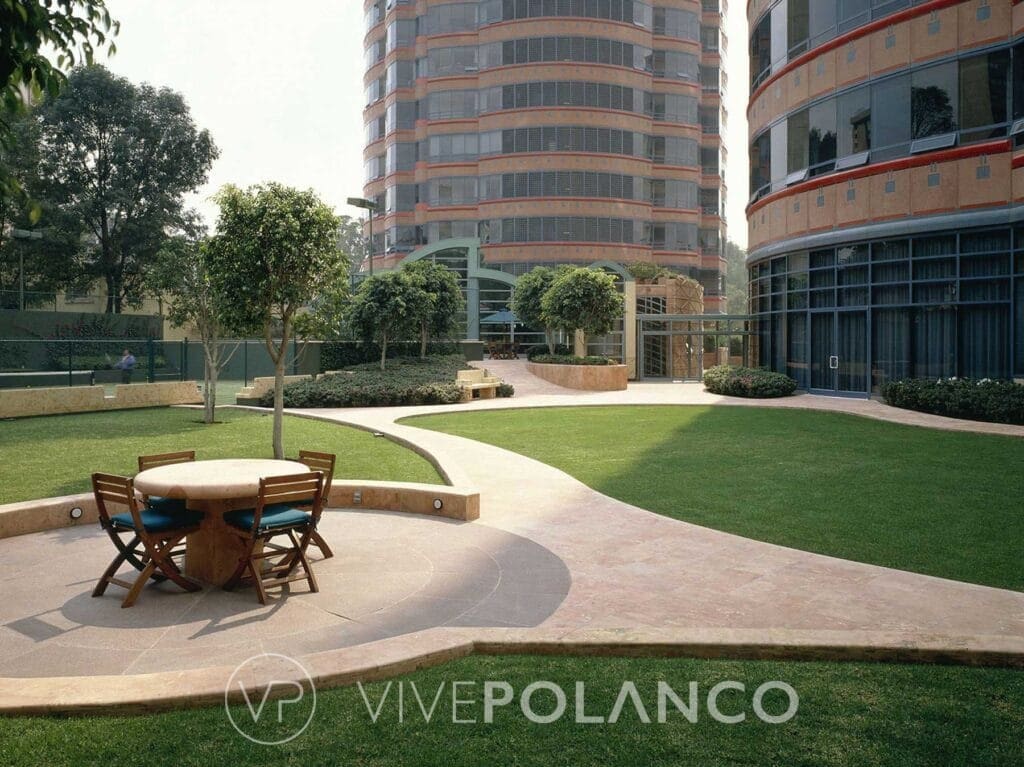 Jardín con mesa y sillas de madera, senderos pavimentados y edificios residenciales modernos