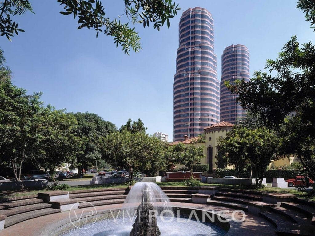 Parque público con fuente y escalinatas, rodeado de vegetación con torres residenciales al fondo