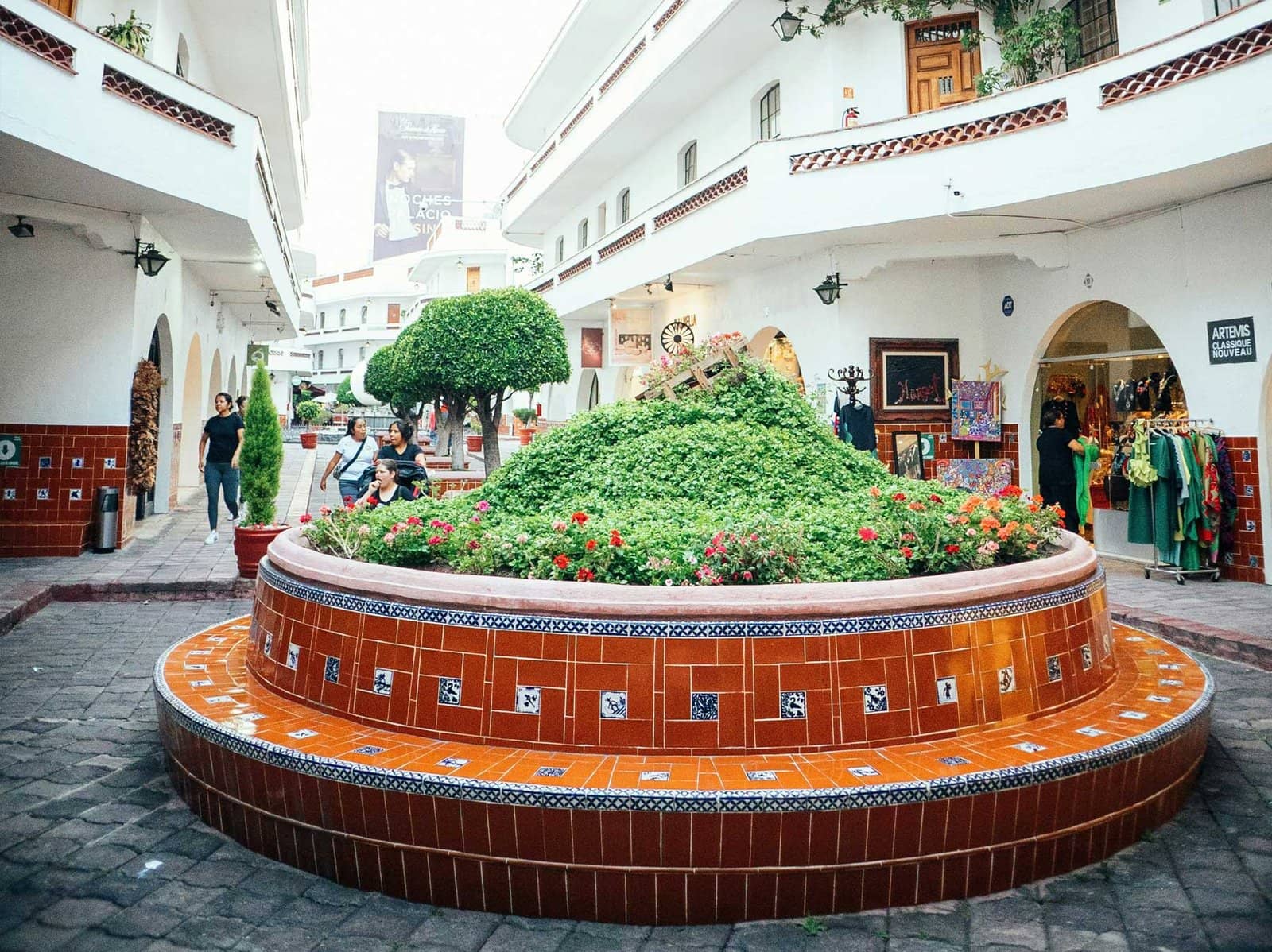 Patio interior de estilo colonial con jardinera circular de ladrillos y azulejos, rodeada de locales comerciales en Pasaje Polanco, Polanco