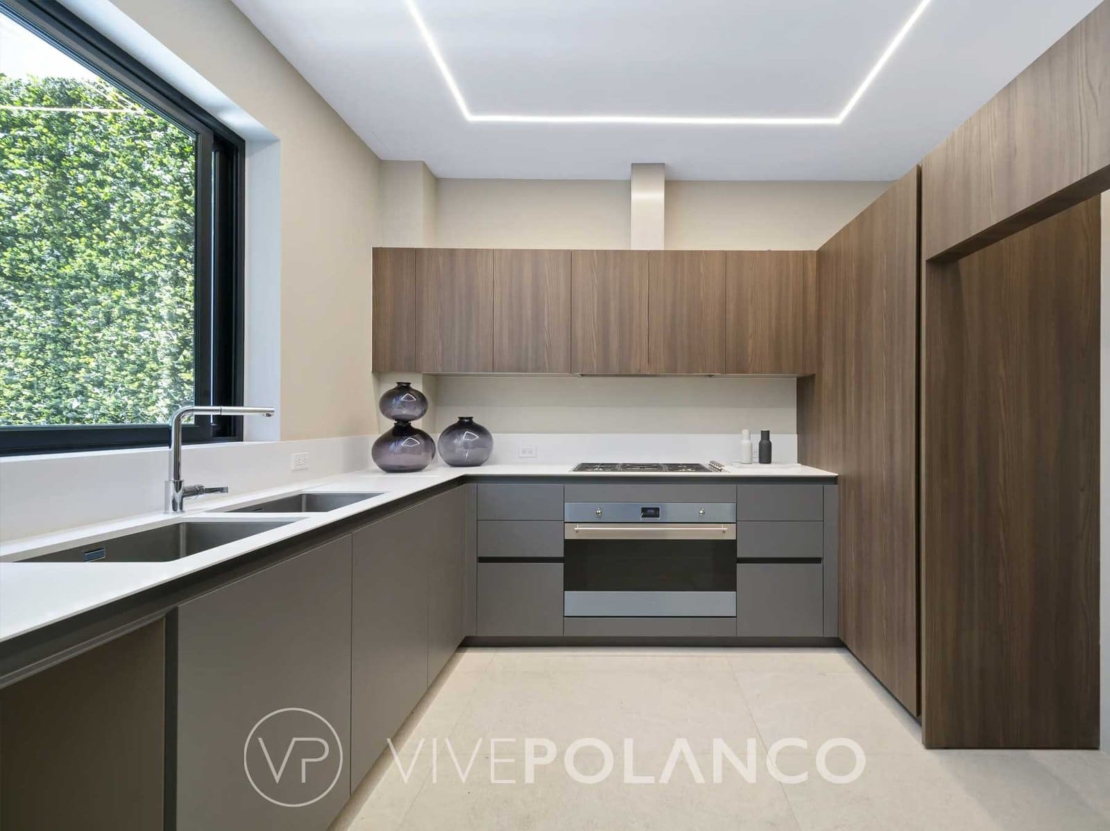 Cocina en L con muebles de madera y gris, parrilla, horno empotrado y una ventana amplia hacia un muro verde