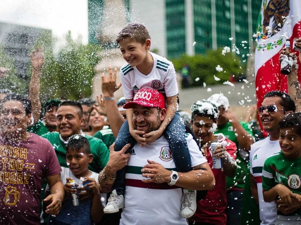 Multitud celebrando con jerseys de la selección mexicana, confeti volando y edificios modernos de fondo